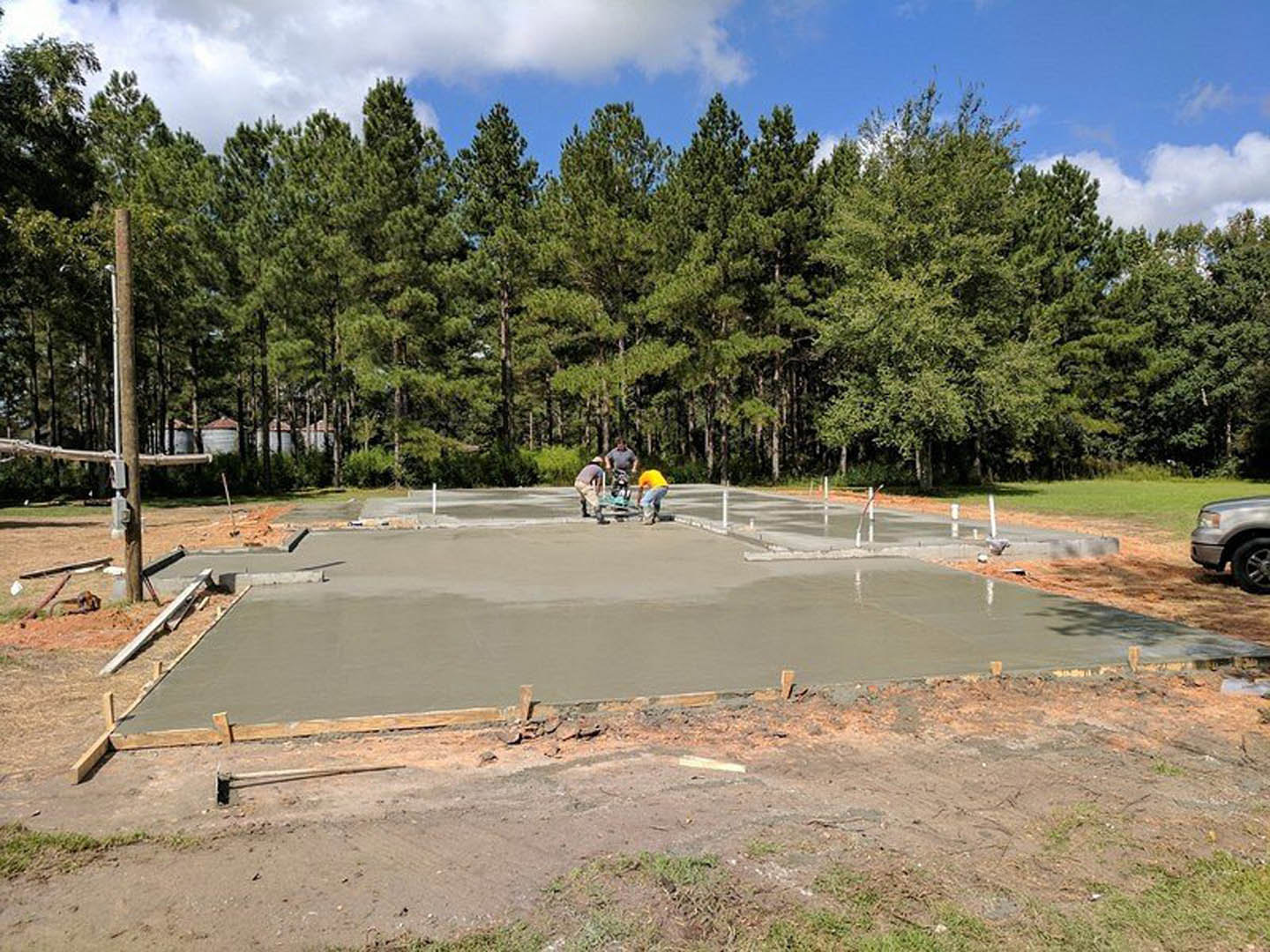 Crew pouring and smoothing concrete slab foundation outdoors, surrounded by grass, trees, and blue sky with clouds