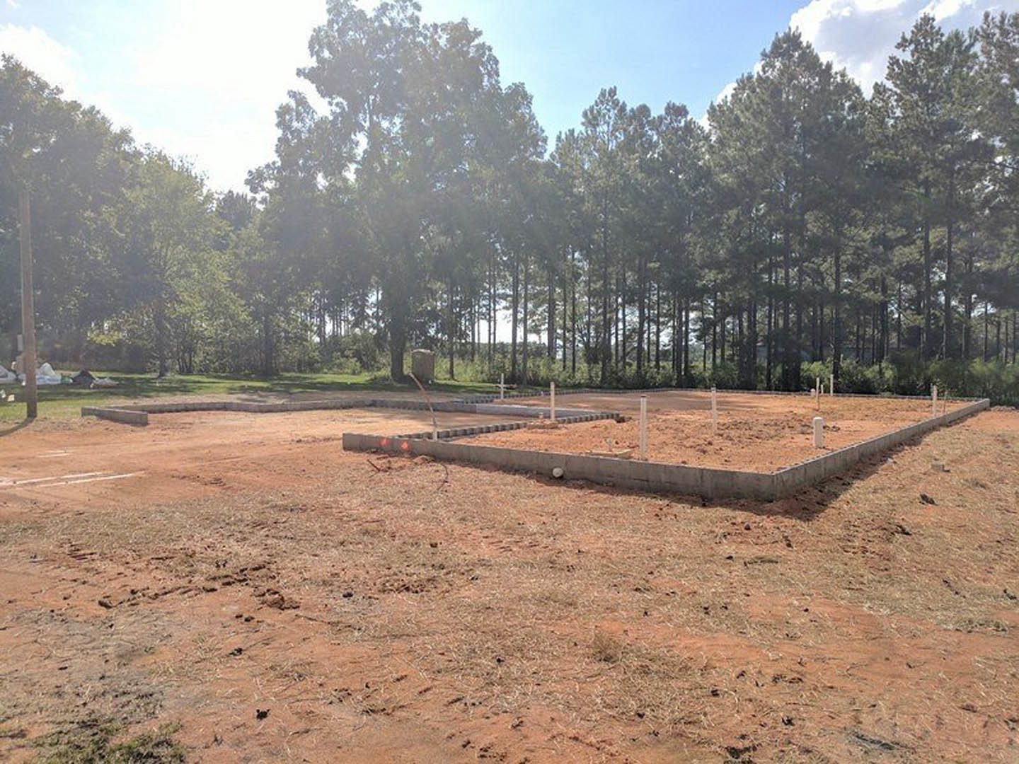Concrete foundation slab surrounded by dirt lot, white marking stakes, and temporary fencing, with mature trees and green foliage in the background