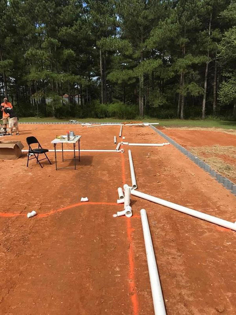 White PVC pipes emerging from clay soil at a residential construction site, with a black folding chair nearby and scattered materials including a brown bag and a table with a can