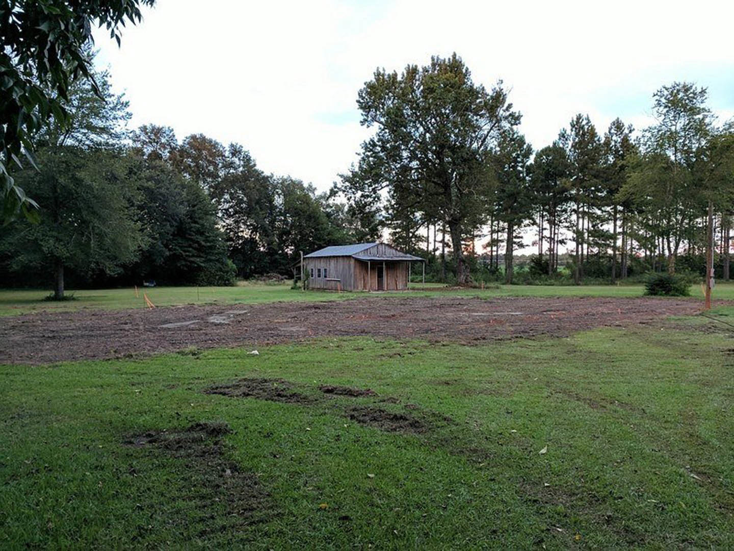 Wooden house with covered porch set on a grassy lawn, surrounded by trees and open field under a cloudy sky
