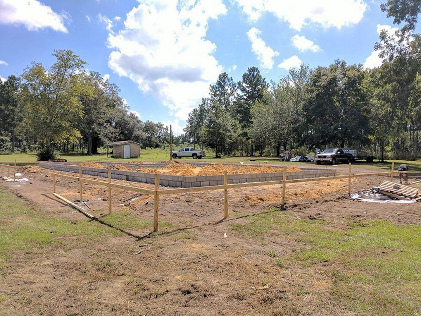 Wooden privacy fence surrounding a grassy construction site with a partially built home, exposed framing, and a small shed under a cloudy sky.