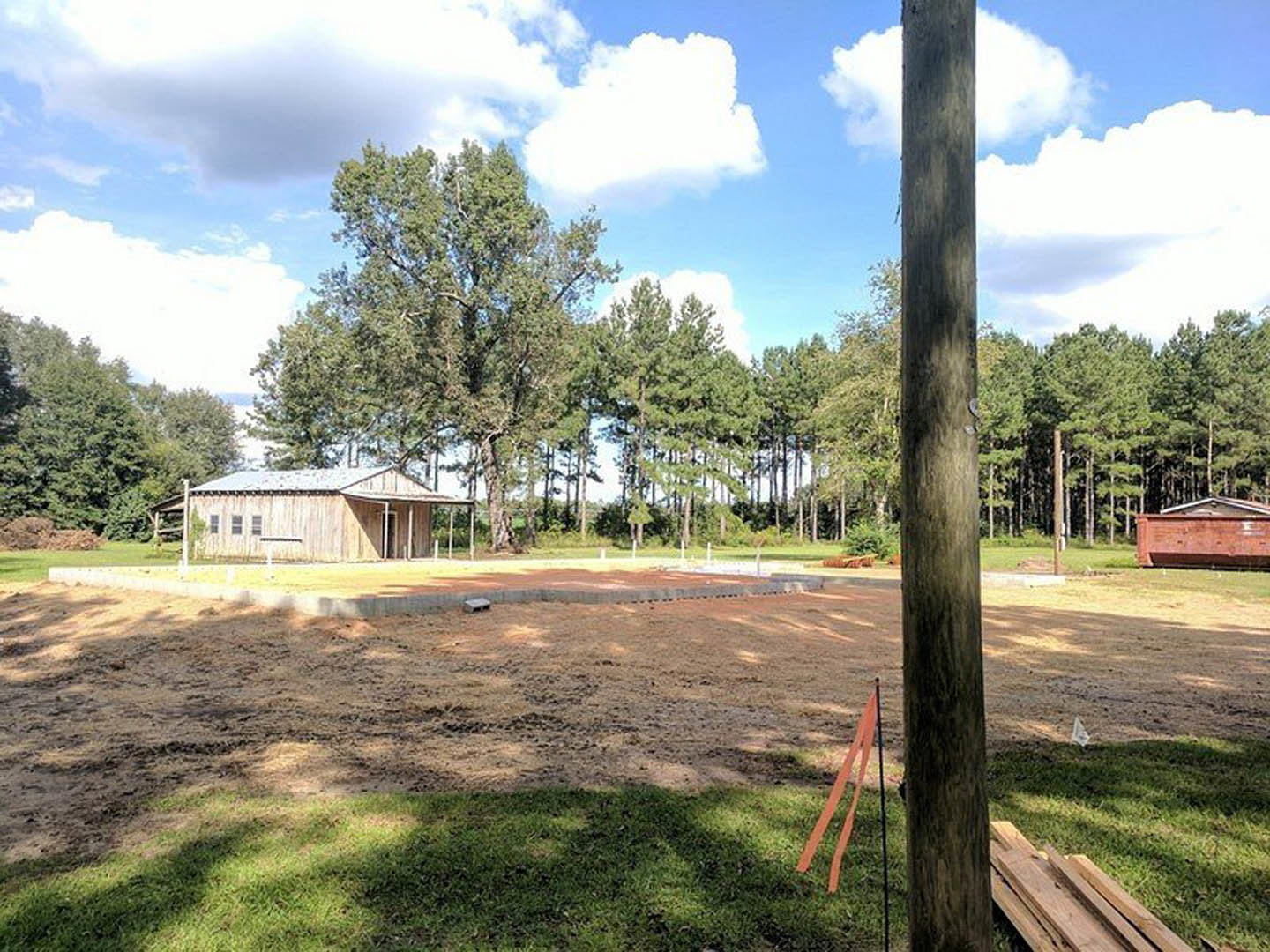 Wood-framed house under construction with red brick walls, exposed roof structure, and piles of lumber on grassy lot surrounded by tall trees.