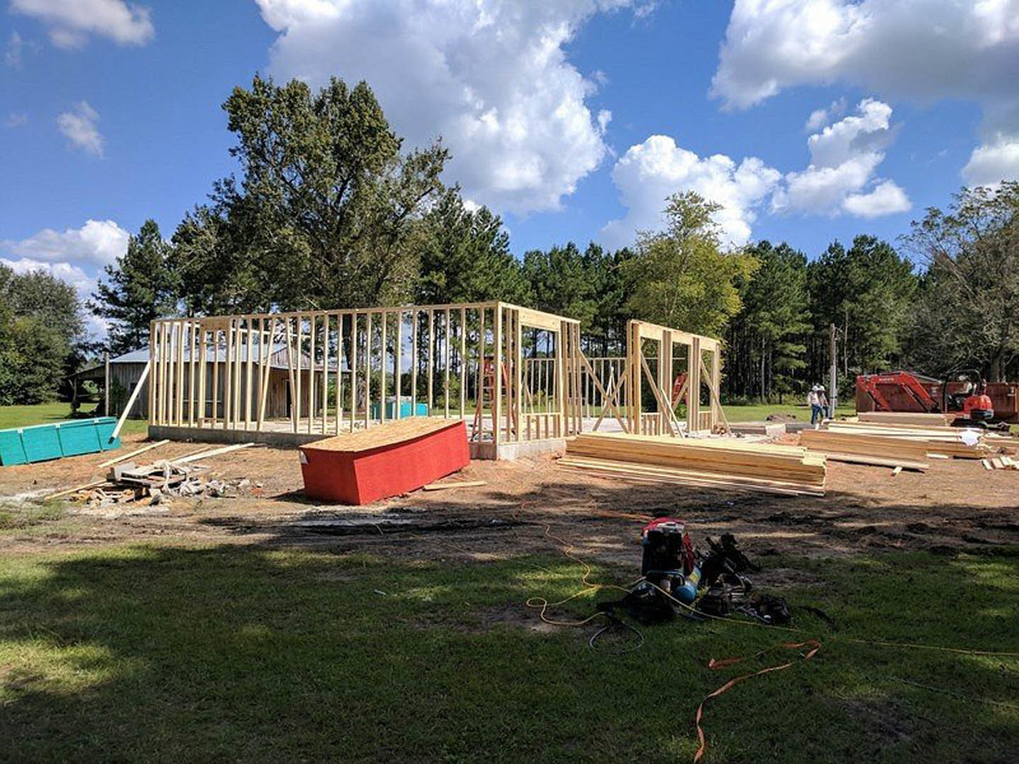 Partially constructed house framed with wood, red exterior wall, wooden fence, grassy yard, blue sky with scattered clouds, trees in background
