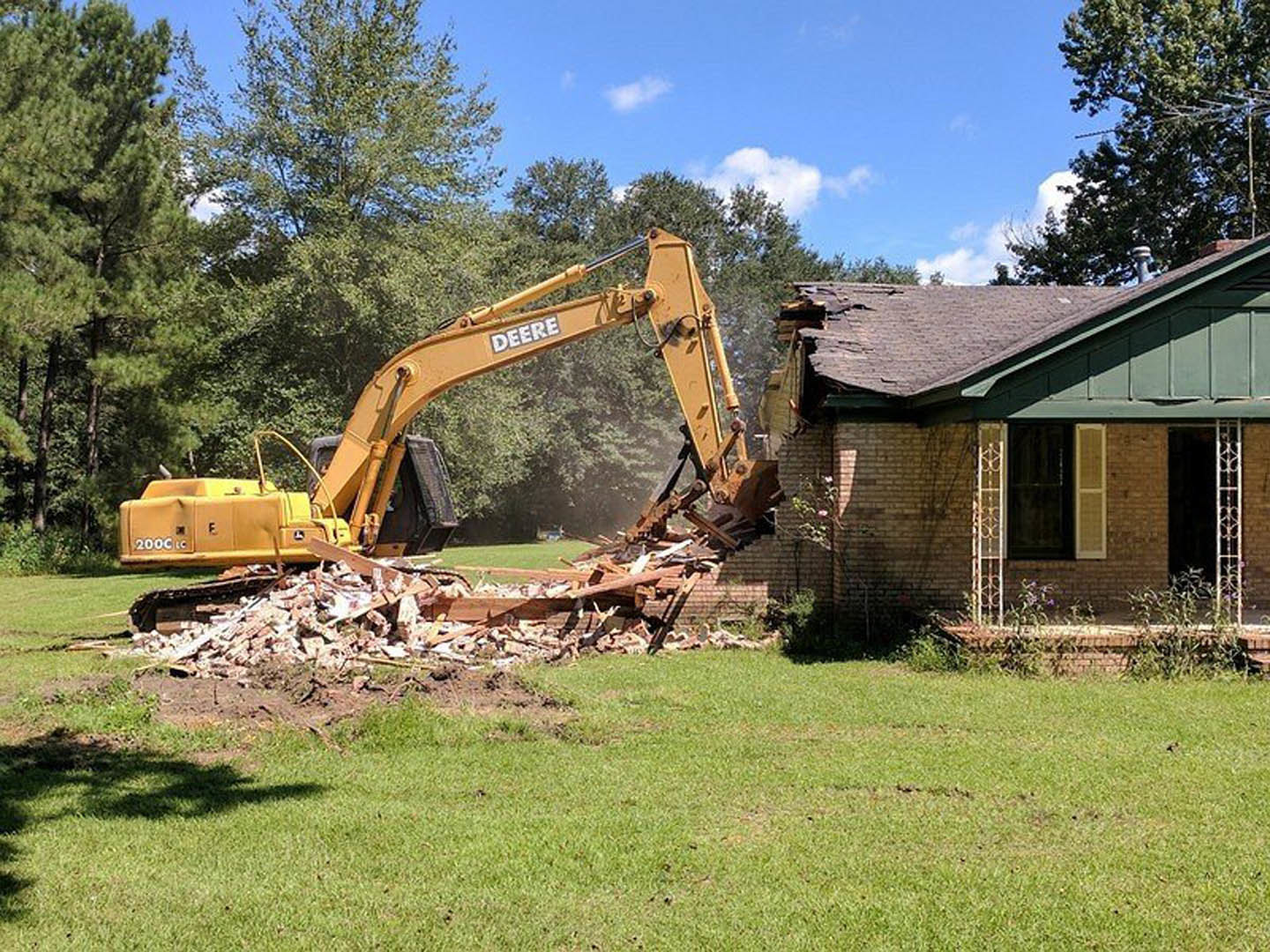 Yellow bulldozer excavating near a house with a damaged roof, surrounded by green grass, trees, and a partly cloudy blue sky