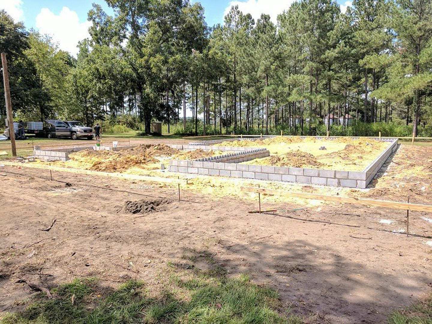 Partially built custom home framed with exposed wood, surrounded by dirt lot, grass, and trees; several people standing near construction site.