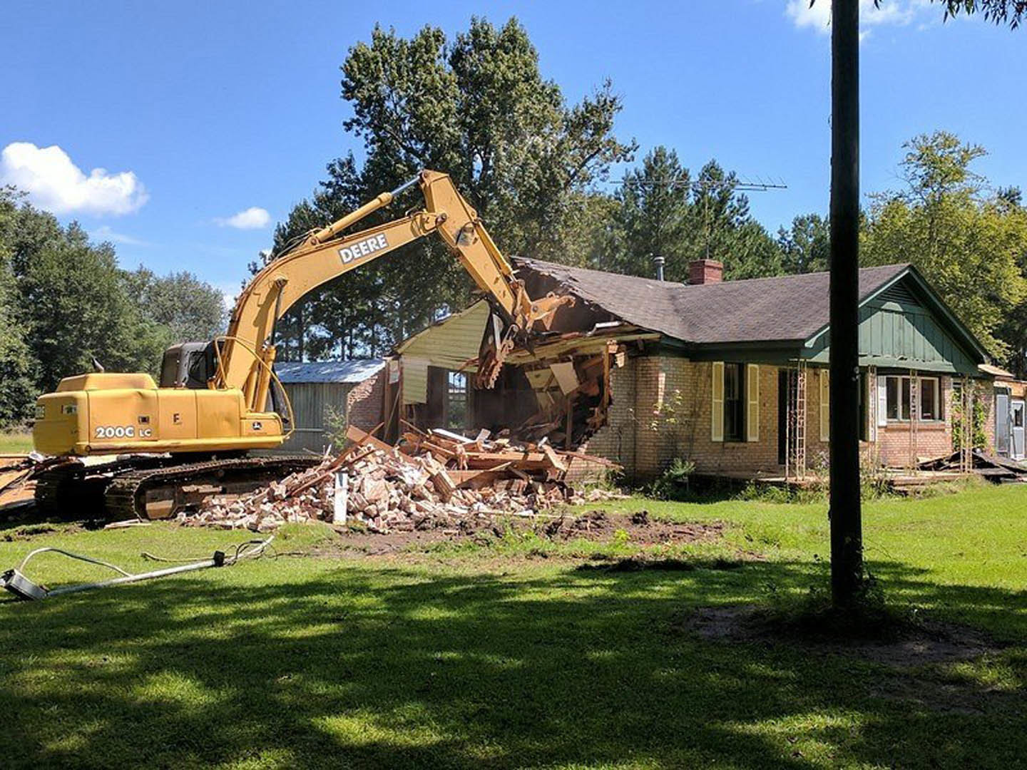 Yellow bulldozer with large bucket demolishing house exterior, broken window visible, green grass field and tree shadow in foreground, blue sky overhead
