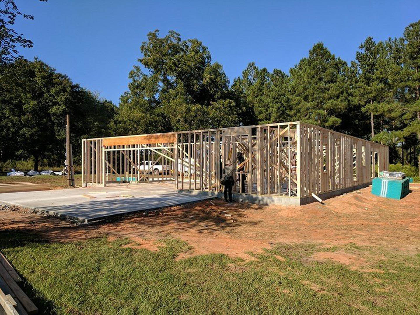 Framed custom home under construction with exposed wood beams, surrounded by grass and mature trees, worker on site, blue storage container nearby