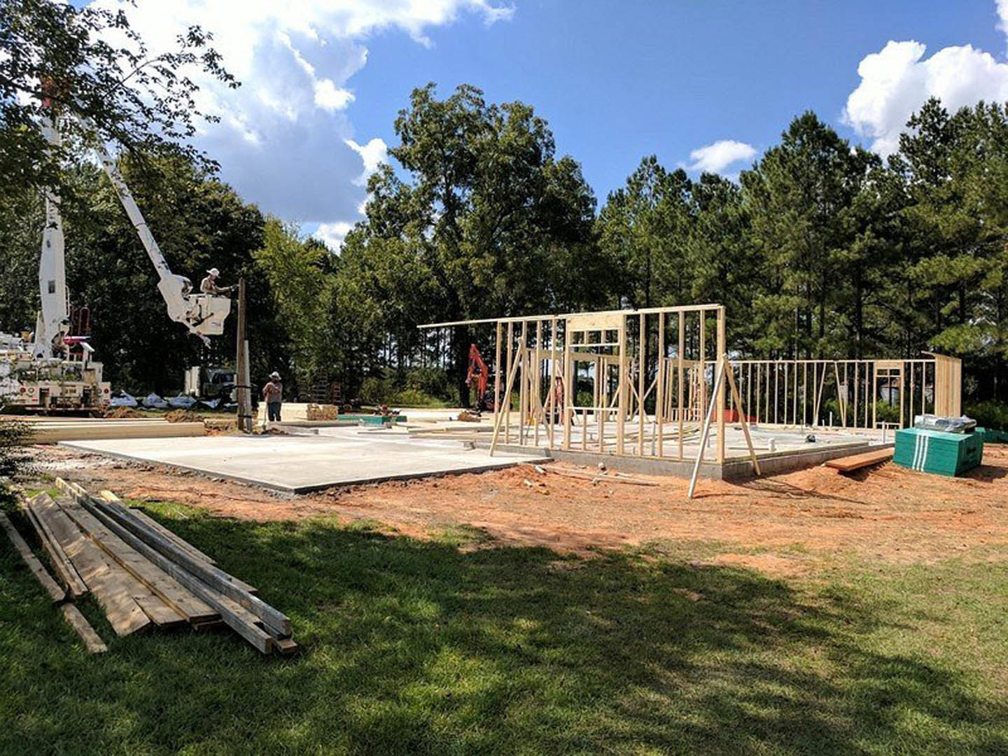 Wood-framed house under construction with crane, pile of lumber on grass, green utility box, concrete fence, and cloudy sky.