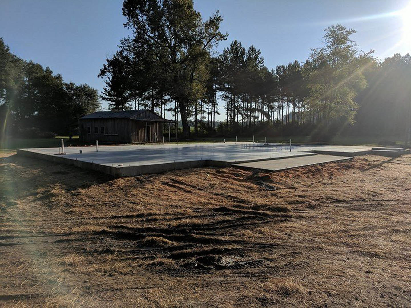 Concrete foundation slab set in a grassy field, bordered by trees under morning sunlight, with a dirt patch and tire tracks nearby, a wooden structure and fence in the distance