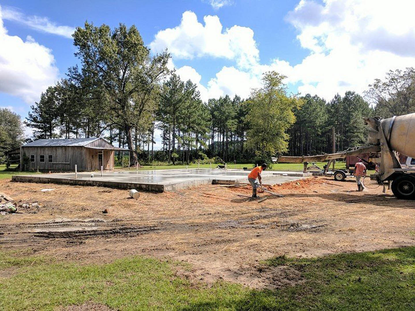 Wood-framed house under construction with snow-covered roof, two men working near foundation, orange shirt visible, tire close-up in foreground, blue sky and trees in background.