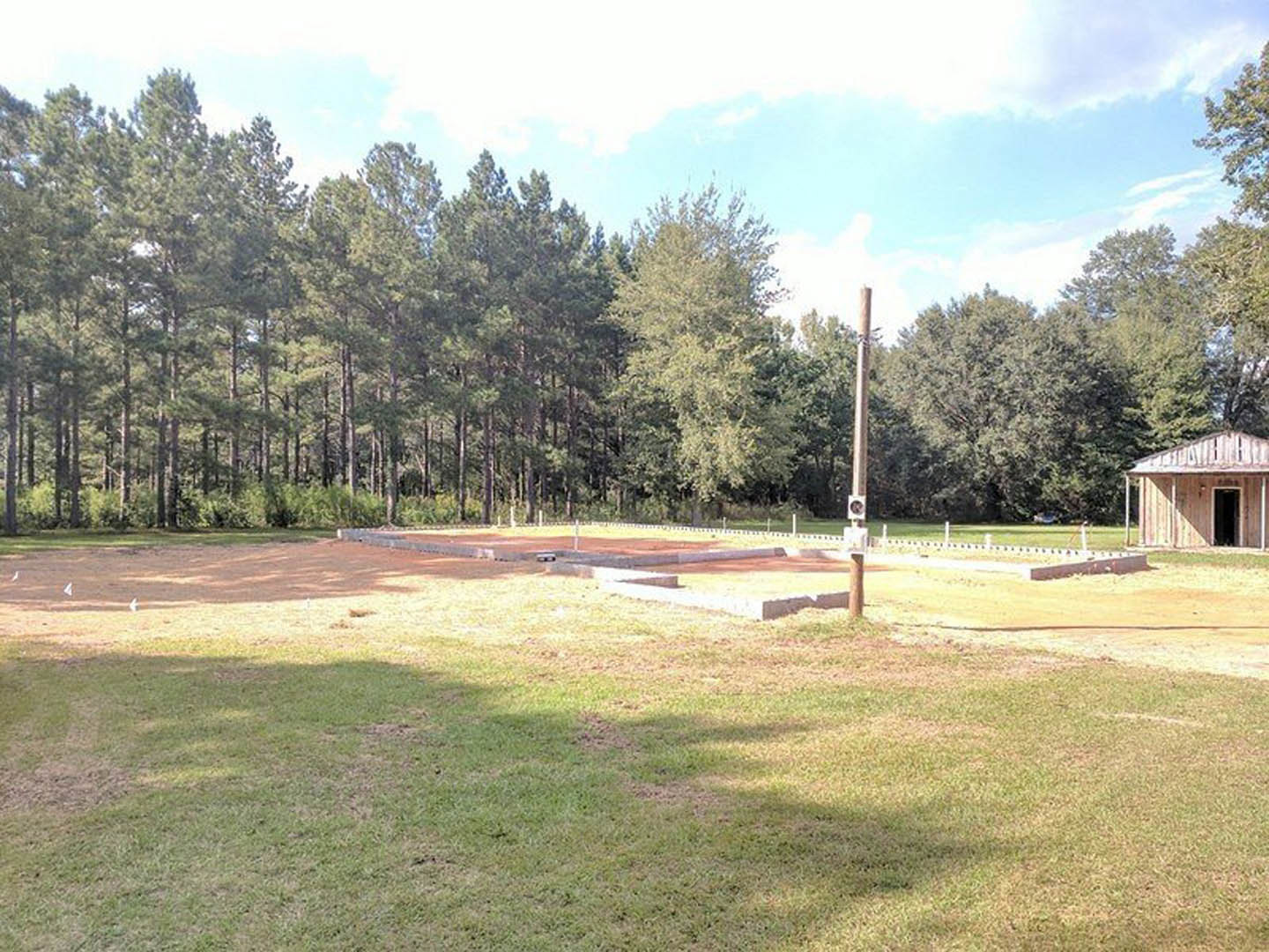 Wood-framed structure under construction on grassy lot, open doorway visible, surrounded by mature trees and cloudy sky