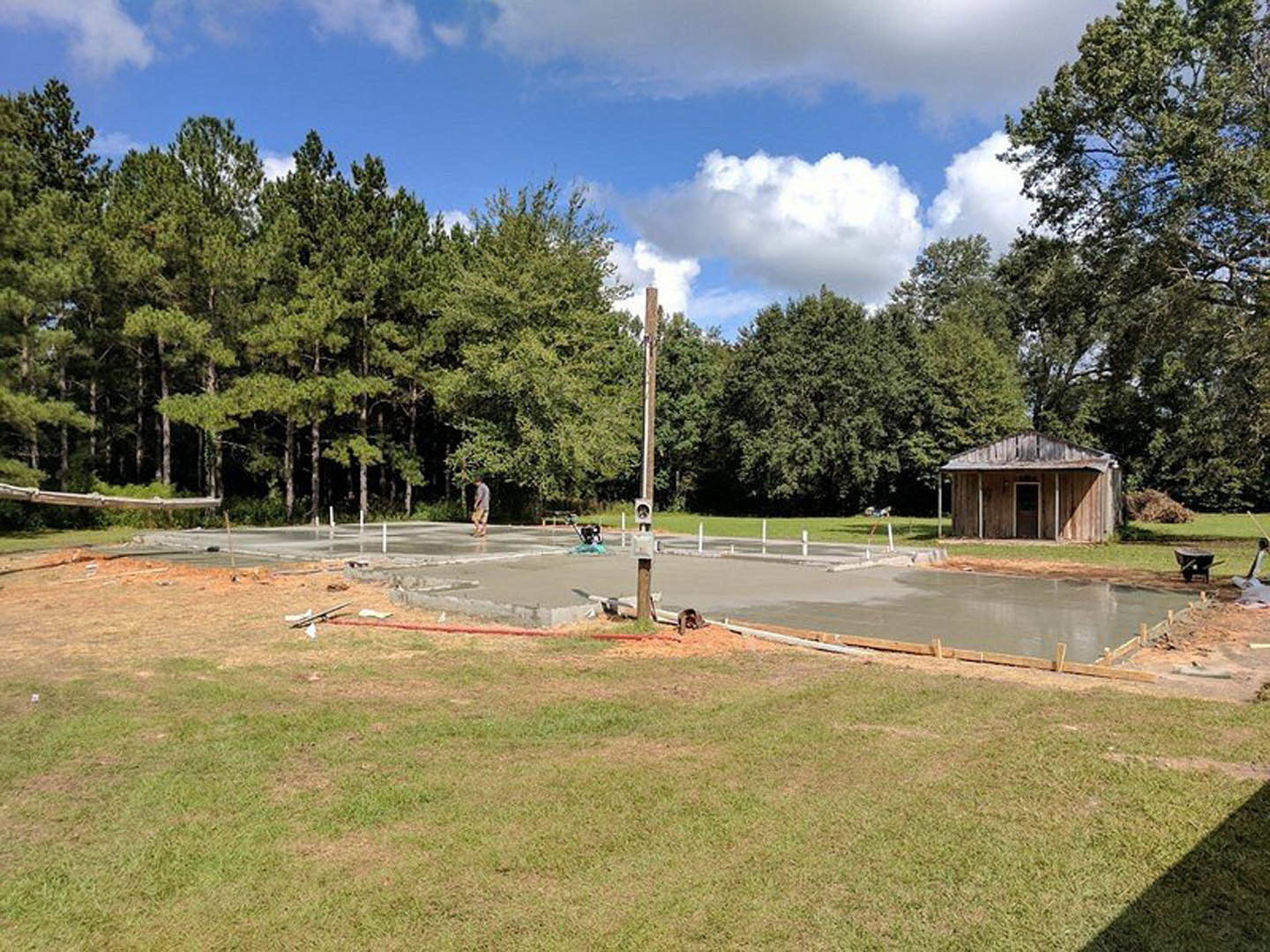 Concrete slab surrounded by grass in a yard, wooden building with door in background, blue sky with scattered clouds, trees and wooden fence along perimeter