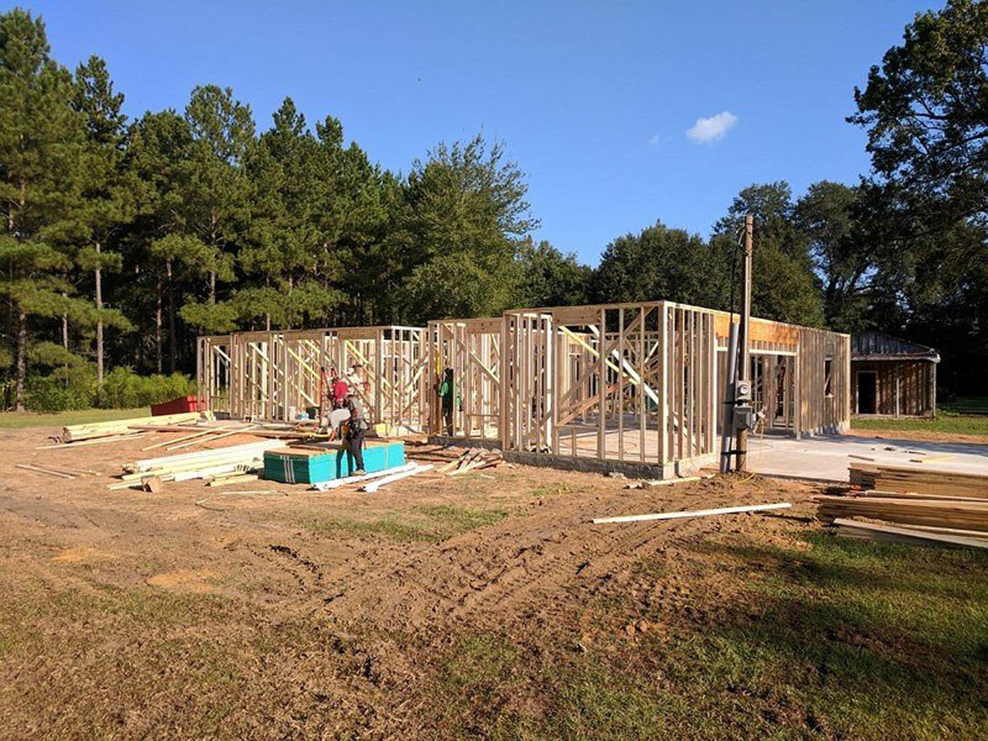 Wood-framed house under construction on a dirt lot, surrounded by trees and blue sky, with construction materials scattered and a person partially visible in the foreground