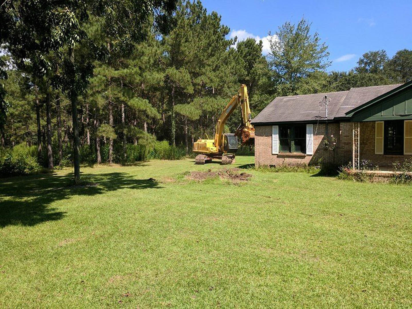 Yellow excavator parked on grassy yard beside cottage-style home with shuttered window, surrounded by trees and open field