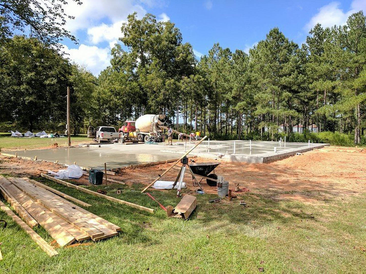 Cement mixer truck parked on dirt construction site surrounded by trees, pile of wood on ground, white truck on roadside, cloudy sky overhead