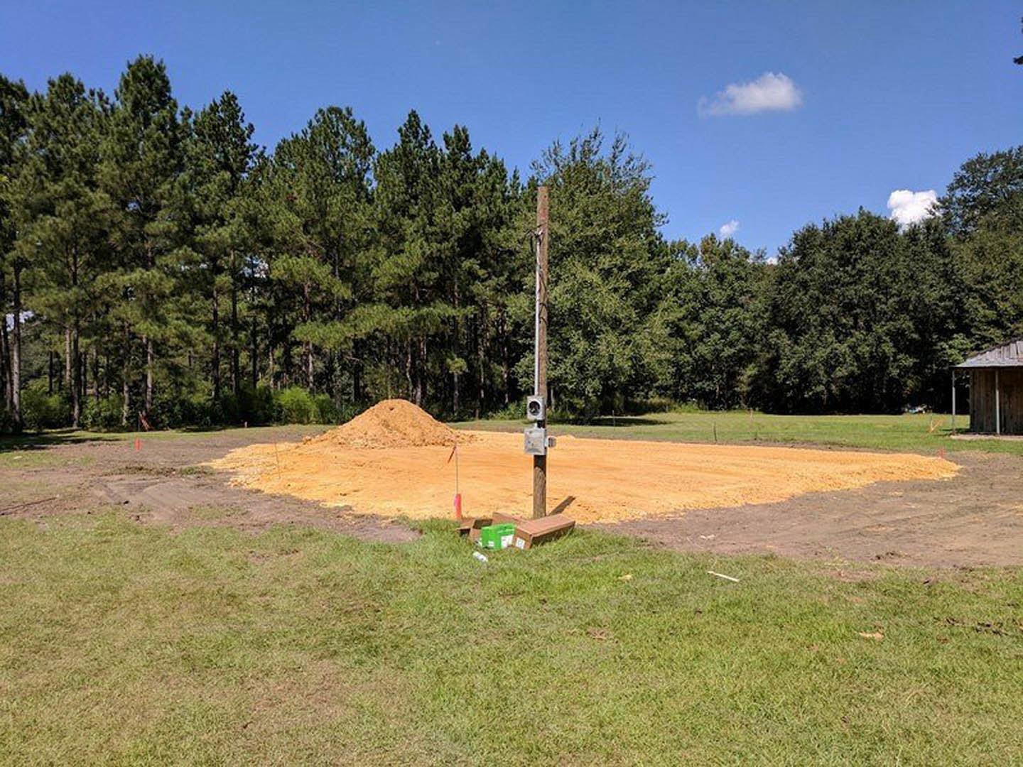 Wooden pole and two dirt piles on grassy field bordered by trees, with Town Creek Indian Mound visible in the background under a partly cloudy sky