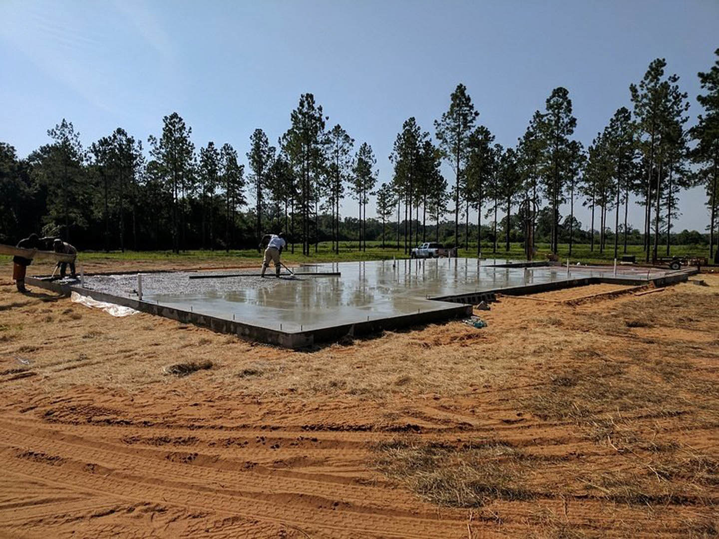 Man in work uniform standing on wet concrete slab with grassy field and trees in background, cloudy sky overhead