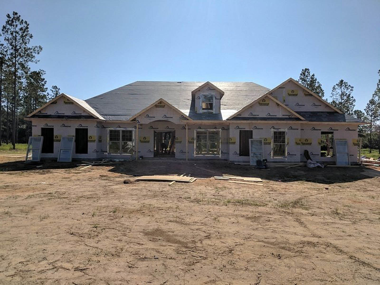 Partially built house with exposed framing and roof, dirt field scattered with wood pieces, man standing in front, trees and blue sky in background, close-up of window visible