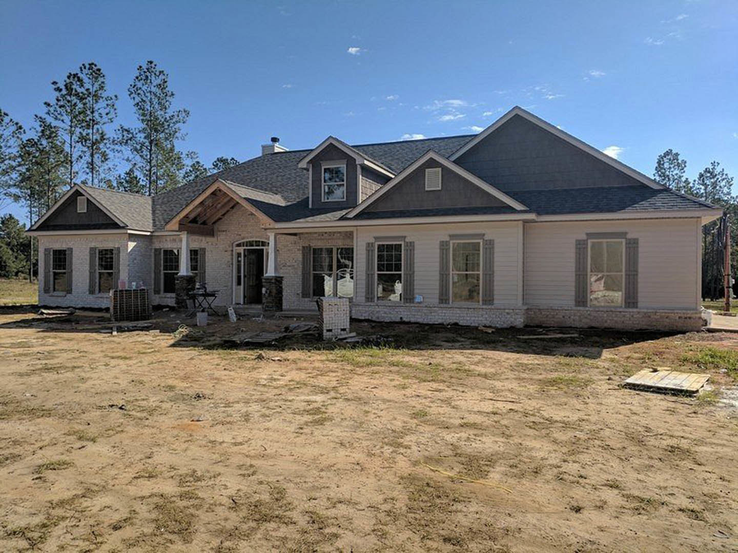 Partially built home with exposed framing, large white-trimmed windows, dirt yard, surrounding trees, and construction truck in foreground