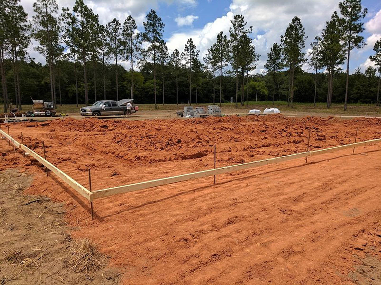 Expansive dirt field bordered by a wooden fence, scattered trees in the background under a cloudy sky, parked car partially visible at the edge