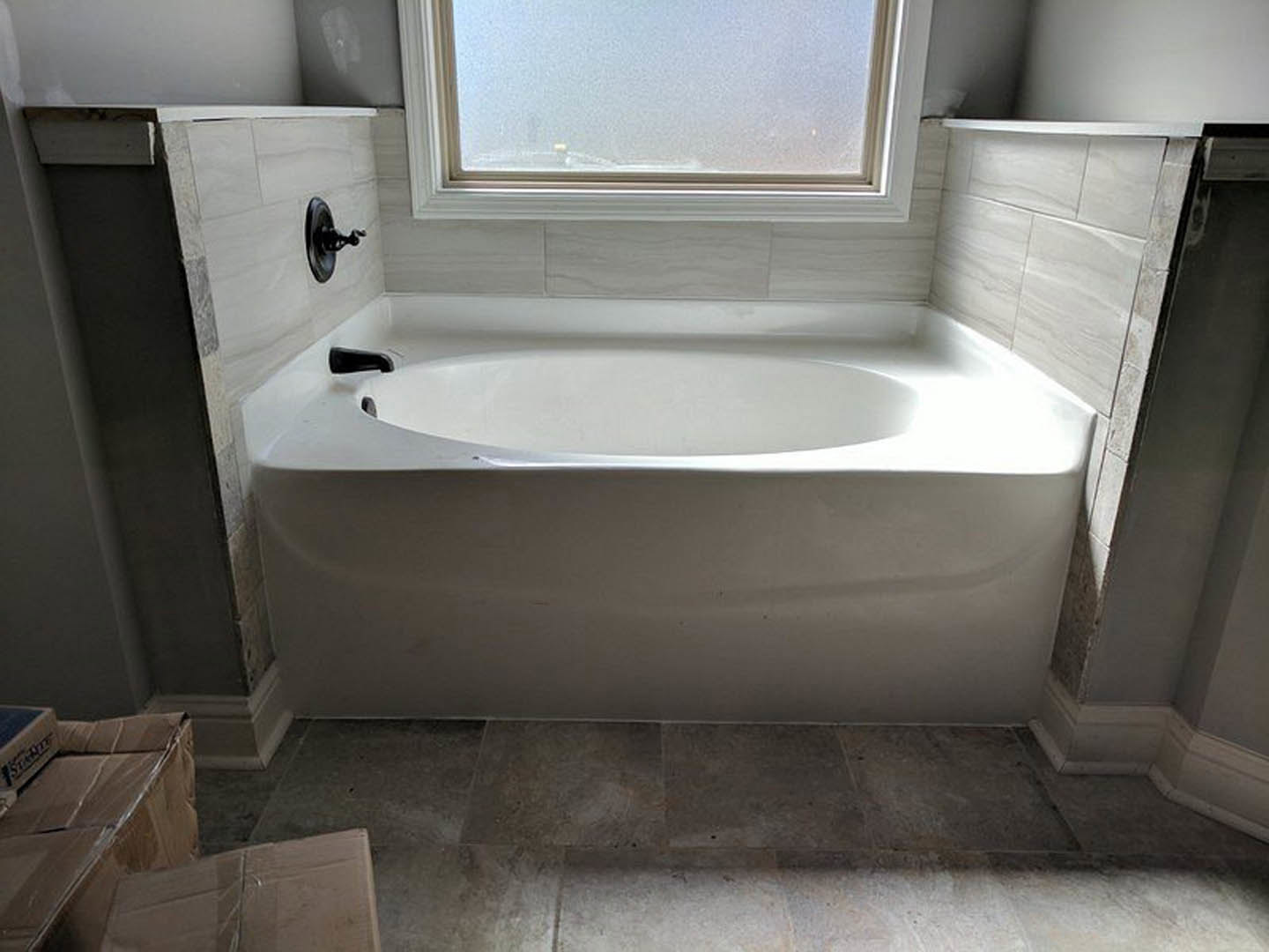 Freestanding white bathtub beside white tile wall, large window with city view, chrome faucet, adjacent sink