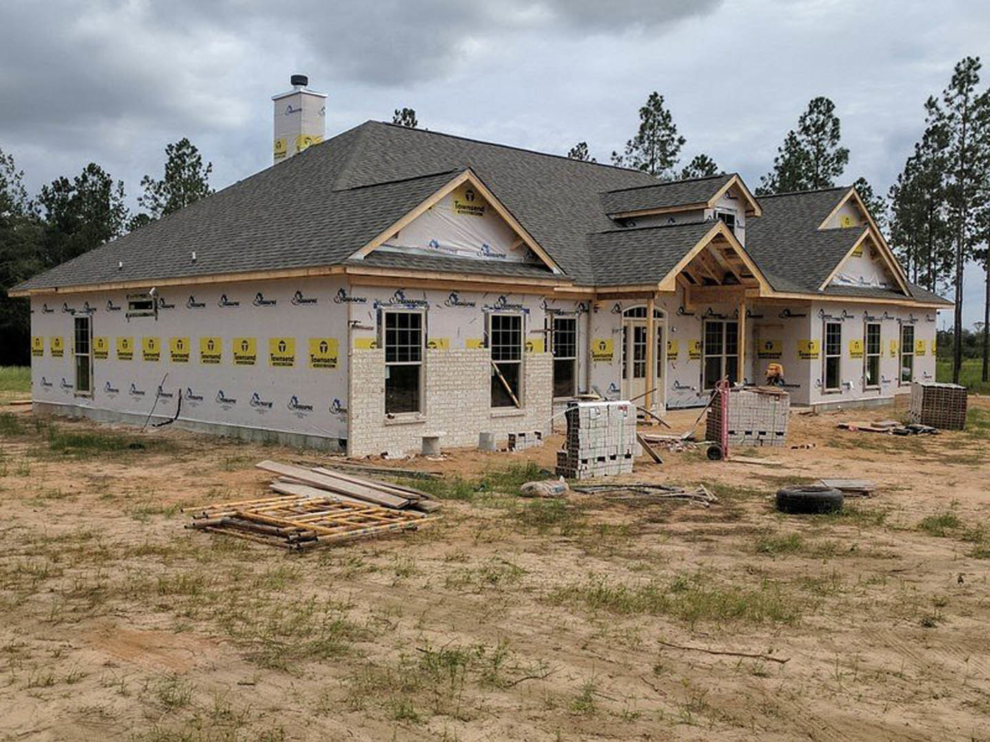 Framed house under construction with exposed wooden beams, stacked lumber, chimney, multi-pane windows, ladder, and pallets on grassy lot