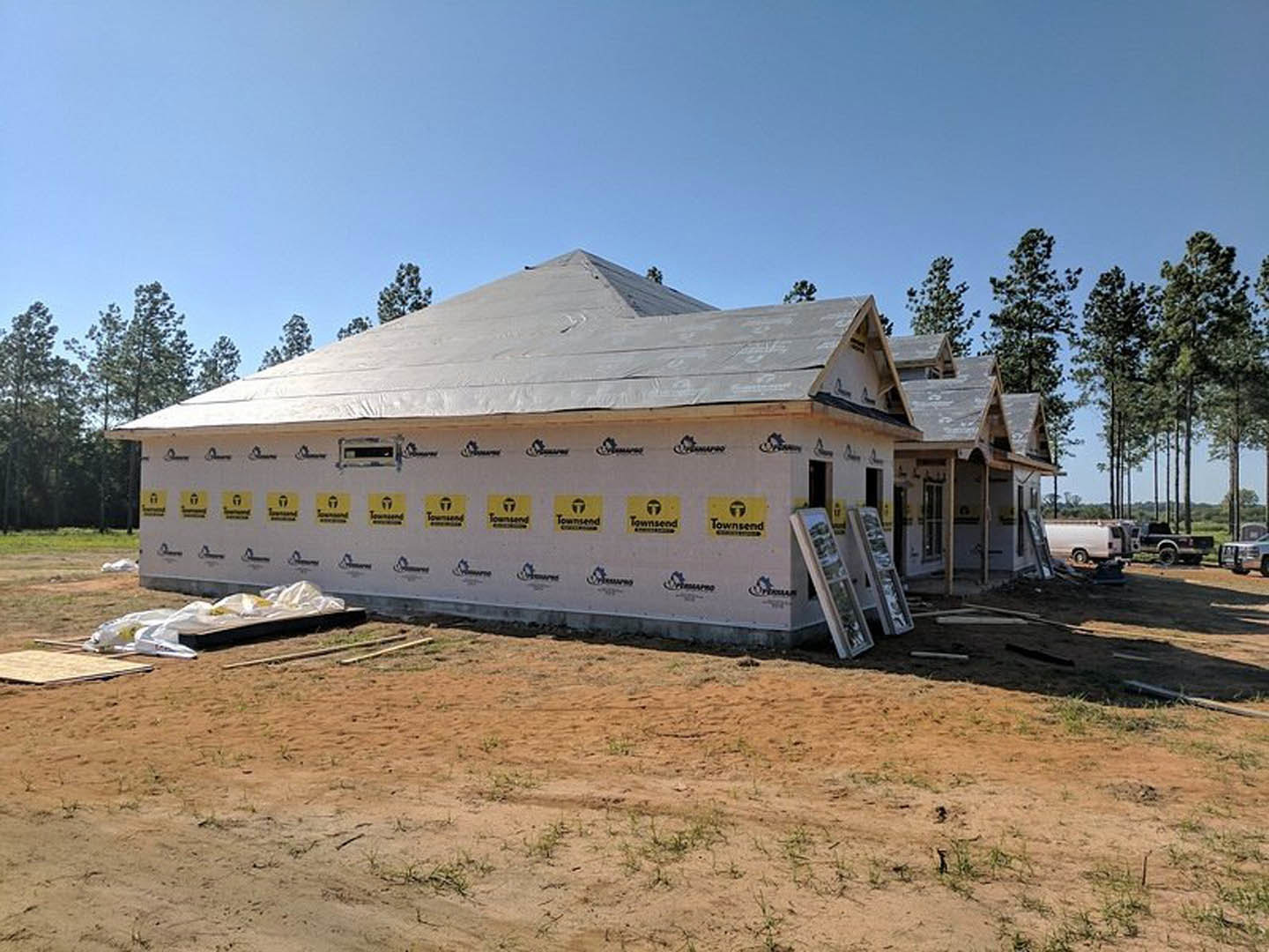 Partially built house with exposed framing and roof, surrounded by dirt lot with patches of grass, several trees nearby, white van parked on site, construction ladder leaning
