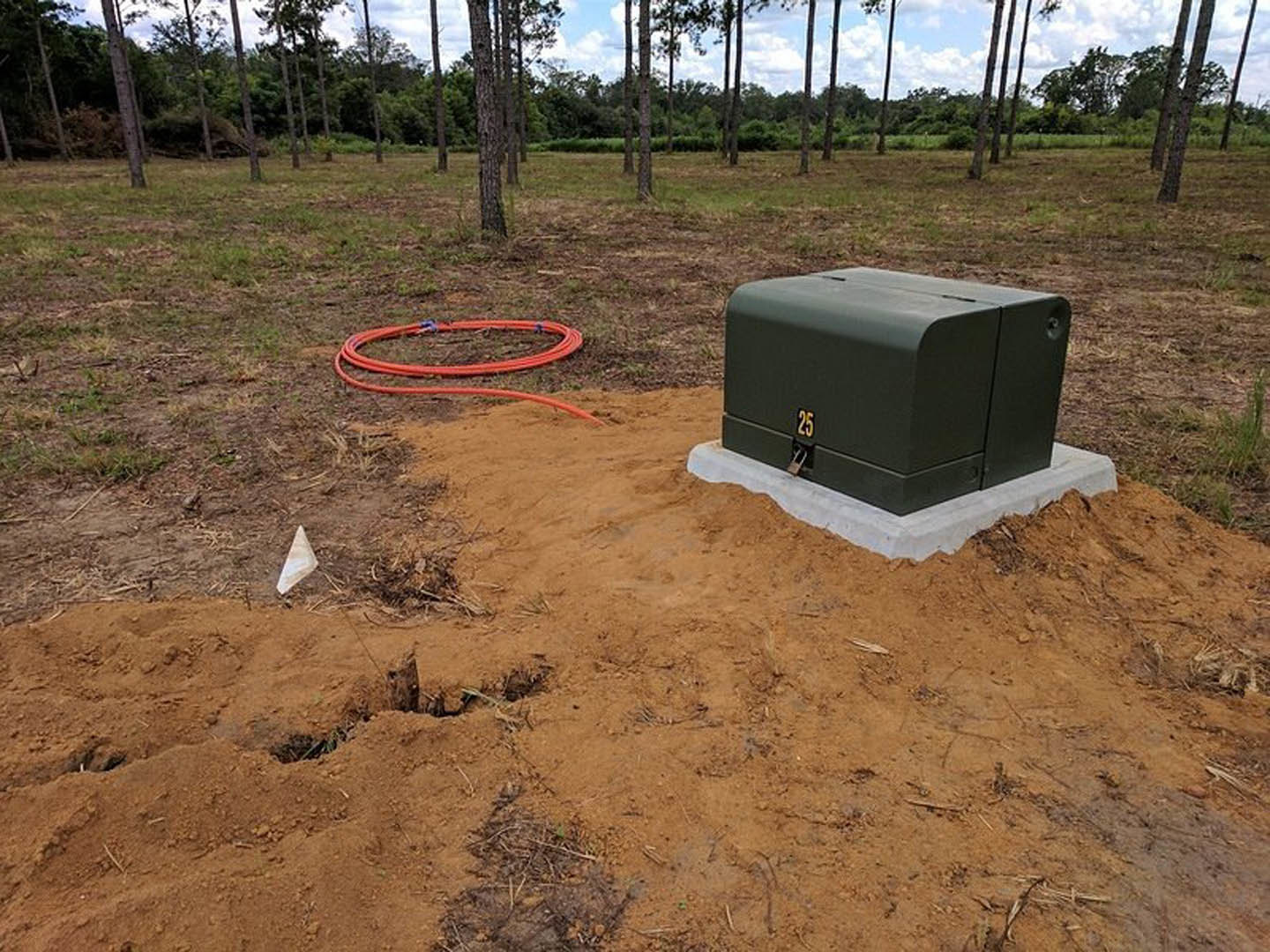 Black metal utility box with lock set in bare dirt near grass and young trees, adjacent to building corner