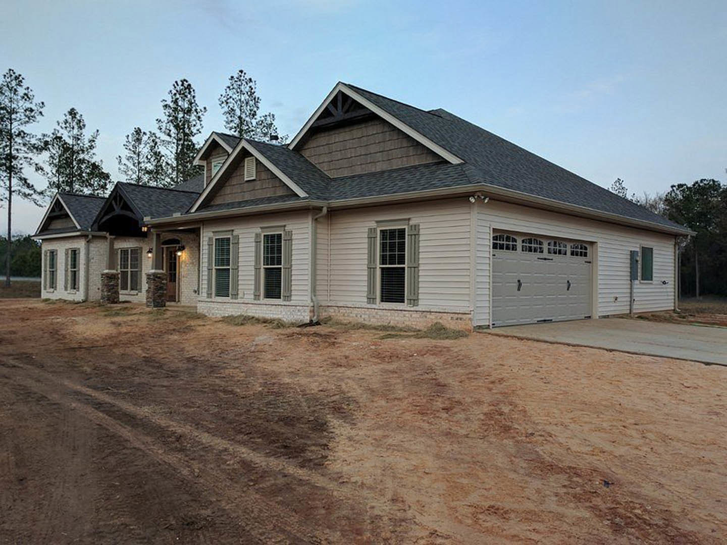 Two-story home with gray siding, white trim, and a shingled roof, featuring a paved driveway leading to a white garage door, front window with dark shutters, and a small dirt patch