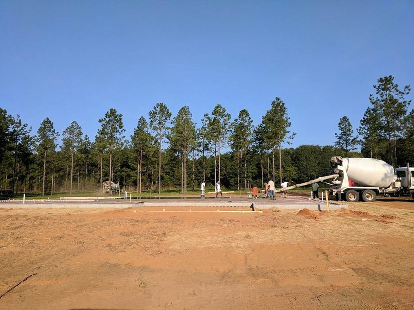 Cement mixer truck parked on dirt field, group of people standing nearby, surrounded by trees under blue sky