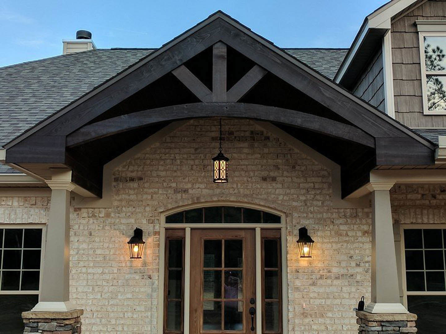 Modern home exterior featuring a wood front door with glass panels, large windows, light-colored siding, and a tree visible through the window.