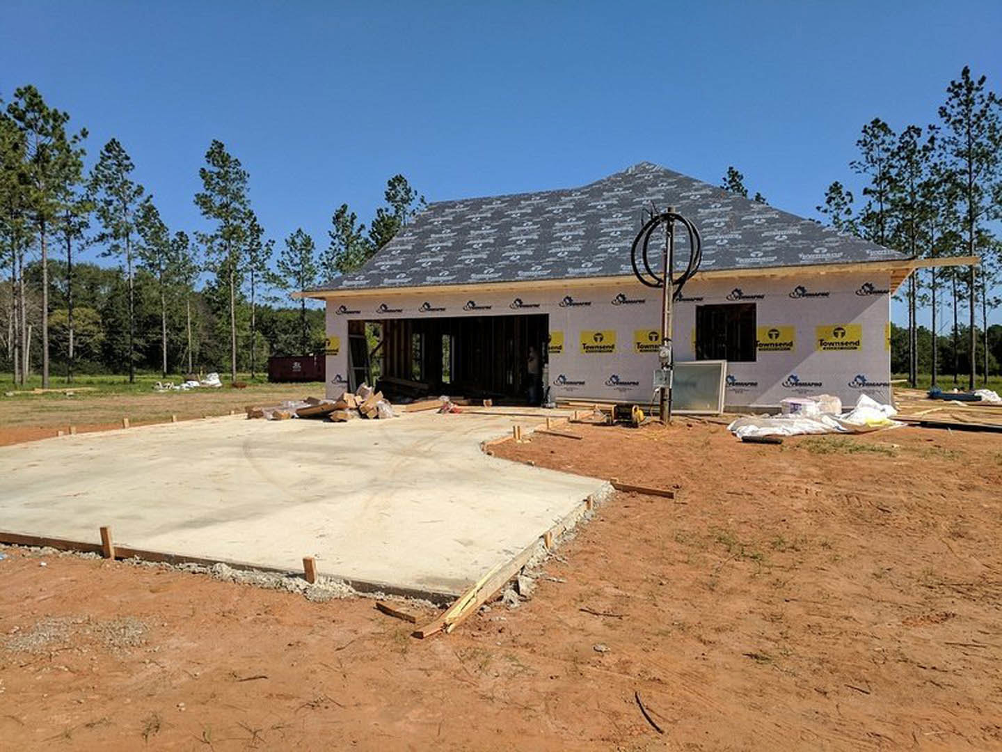 Partially built house with exposed framing, attached garage, unfinished roof, concrete slab foundation, scattered bricks and lumber, visible windows, surrounded by dirt lot and