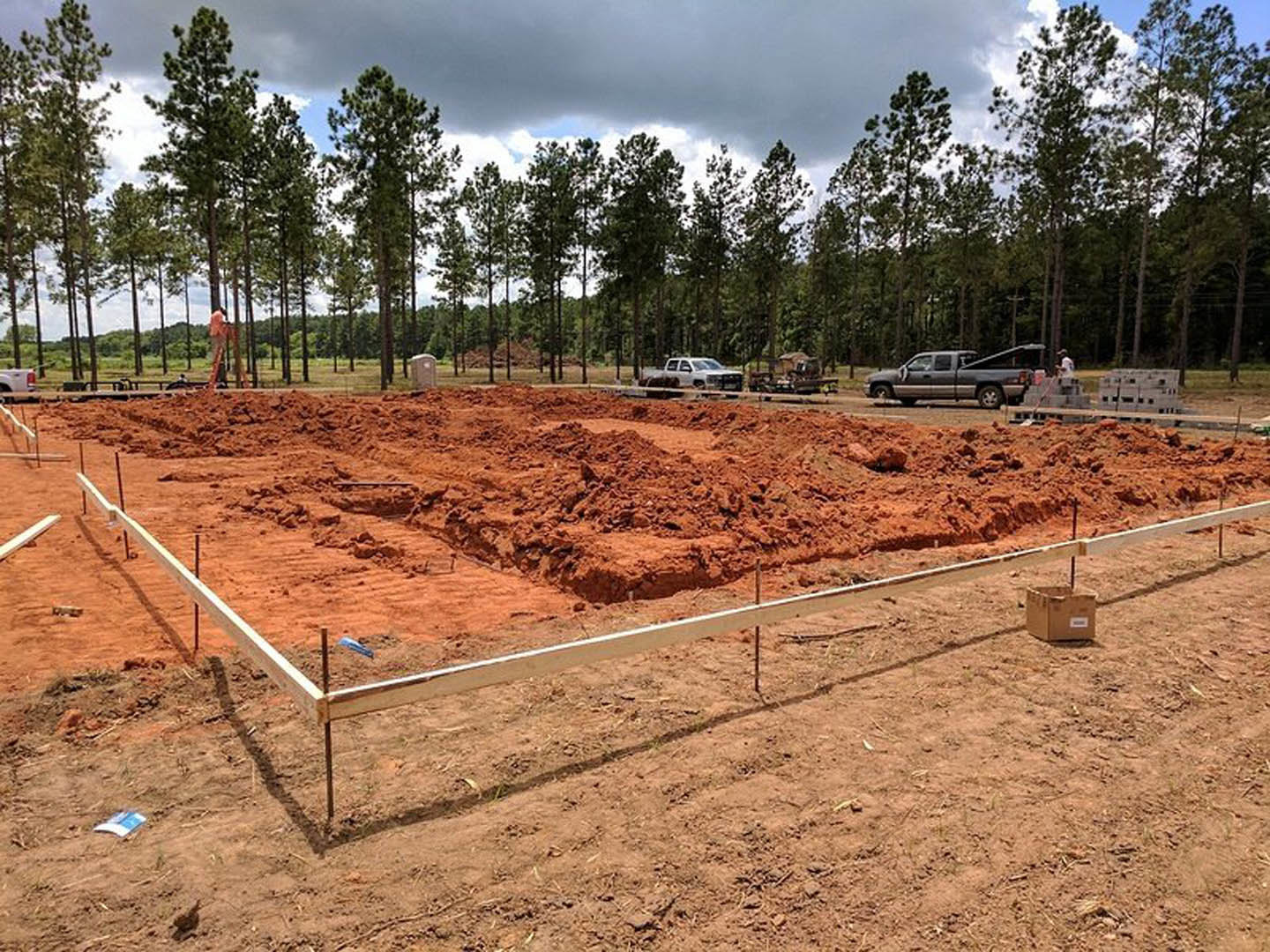 Construction site with dirt piles, brown box on ground, grey truck with open door, fenced perimeter, and row of trees in background