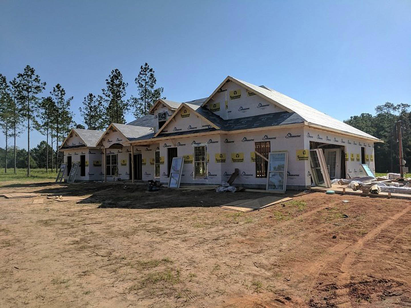 Partially built house with exposed framing, surrounded by dirt field and scattered trees, white-framed window visible on exterior wall
