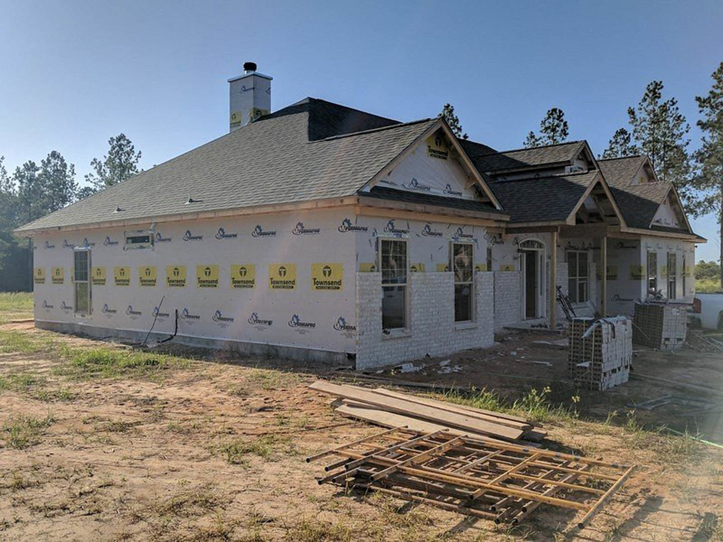 Two-story house under construction with exposed framing, fenced yard, metal scaffolding stacked near the foundation, glass window installed, brick chimney partially built, black