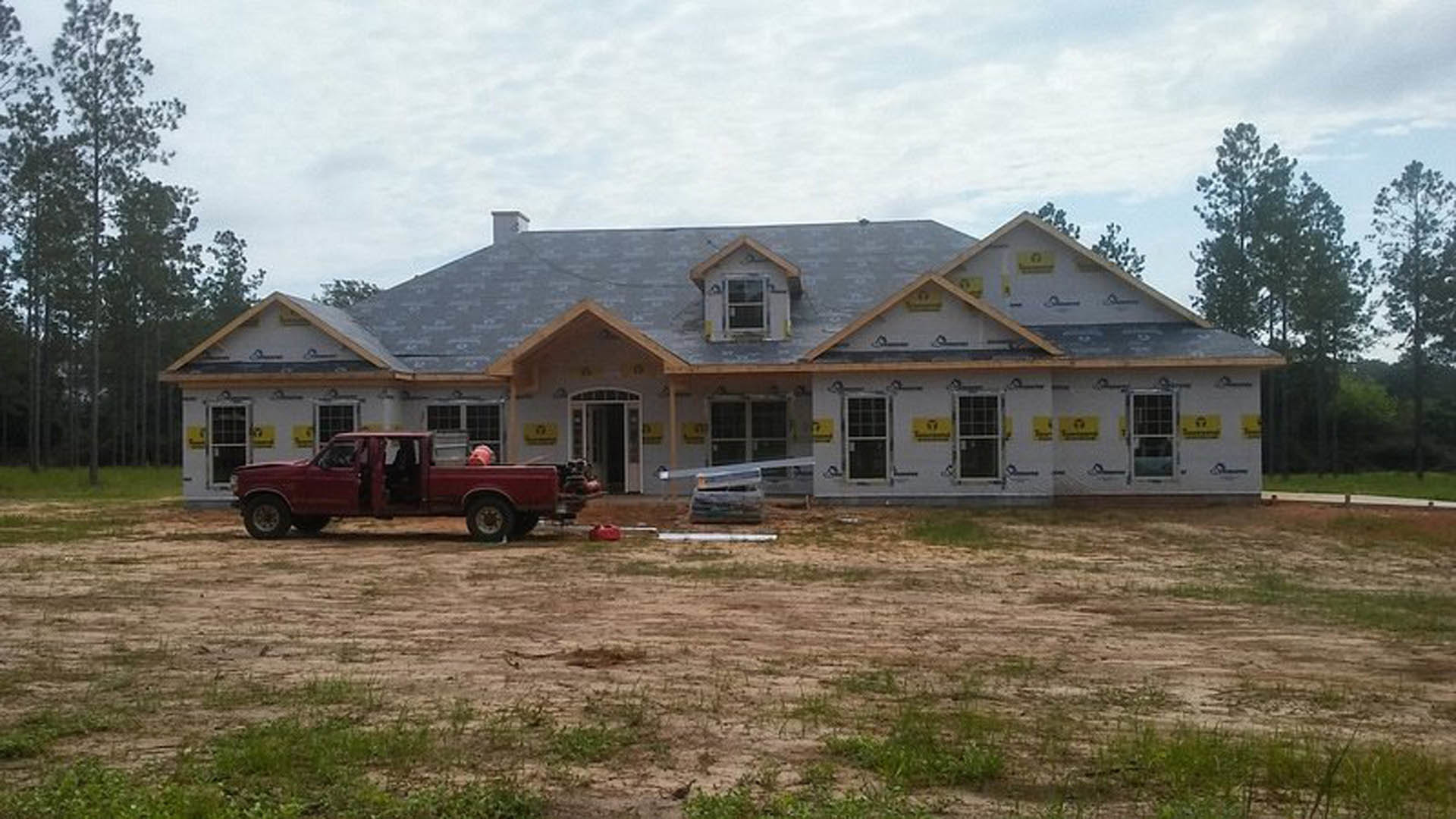 Two-story house under construction with white-framed windows, ladder leaning against exterior, dirt and grass lot, red pickup truck parked in front with open doors, cloudy sky