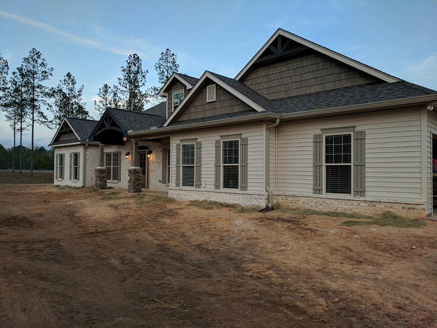 Two-story brick house with white shuttered windows, surrounded by a dirt yard and unpaved road, set against a cloudy sky with trees in the background