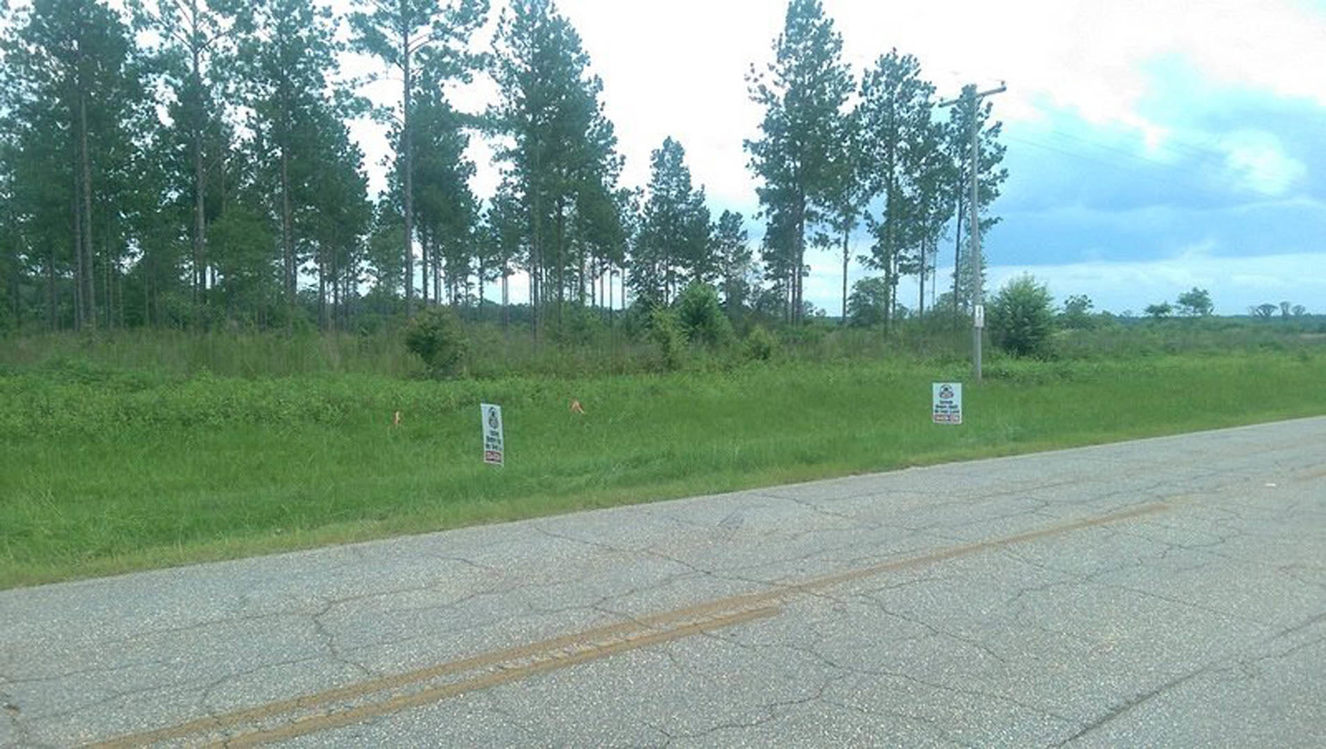 Asphalt road bordered by grass and trees, white sign with black text mounted on tall pole, cloudy sky overhead