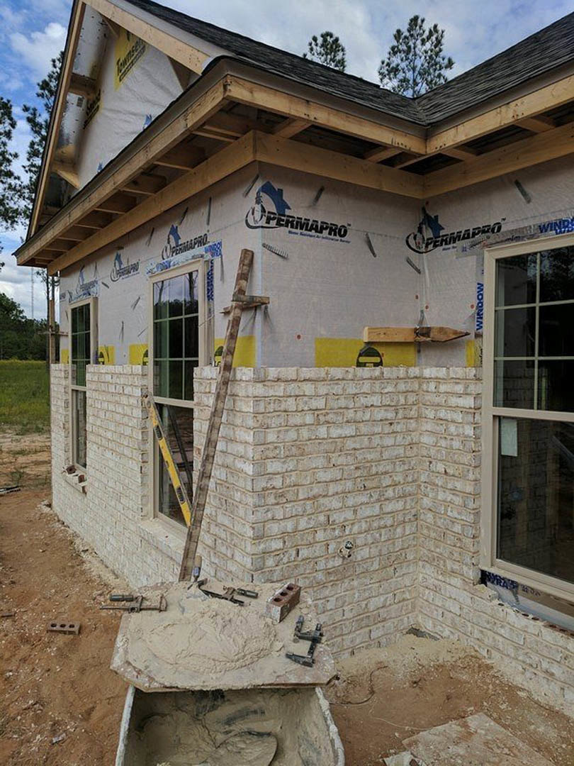 Two-story brick house under construction with scaffolding, wooden ladder, and pile of sand in front; unfinished windows and exposed exterior walls.
