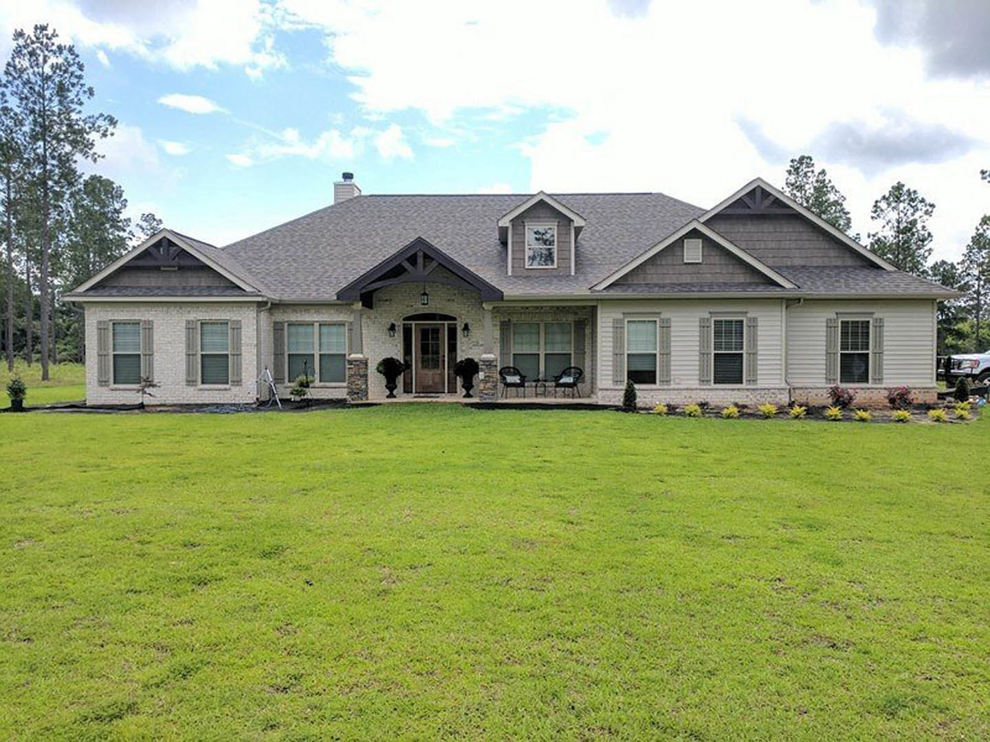 Two-story house with white-framed windows, covered porch with chairs, and manicured green lawn bordered by trees under a partly cloudy sky