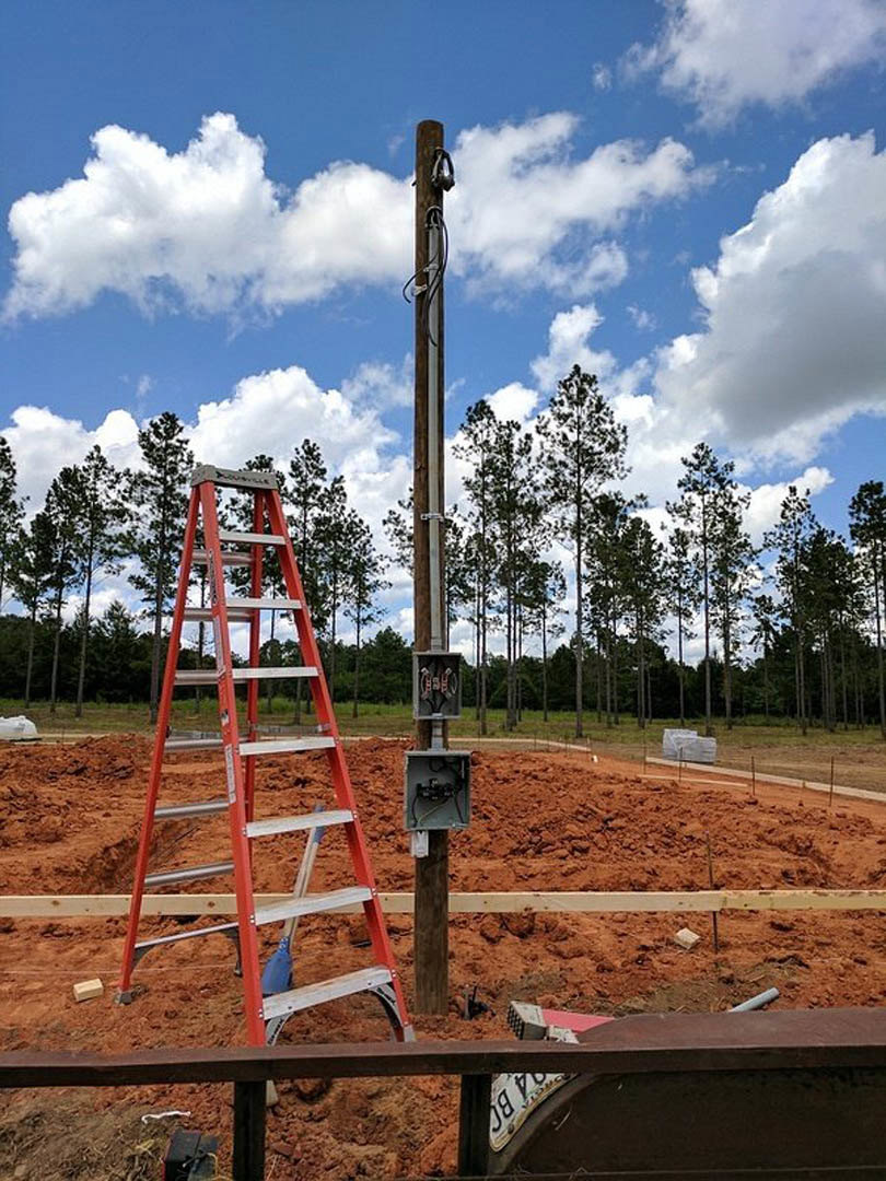 Tall wooden utility pole with metal ladder standing on bare dirt, surrounded by trees and cloudy sky in the background.