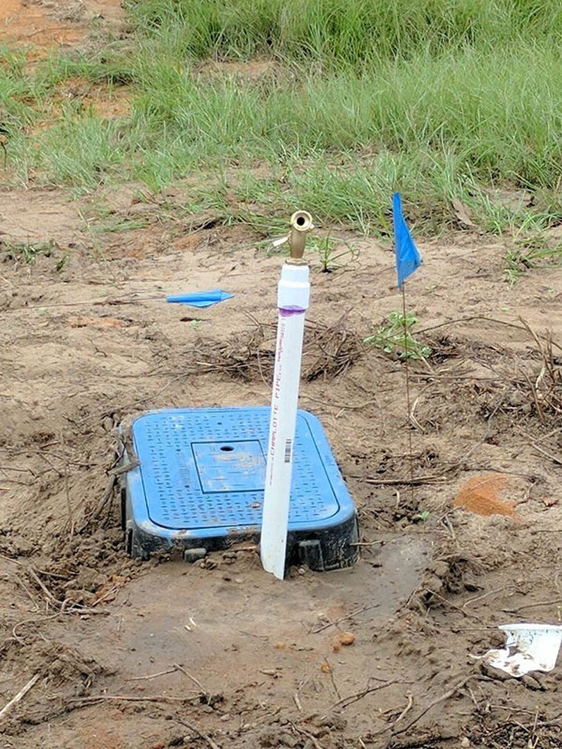 Ground-level water sprinkler with white pipe and red writing, blue square object with white pole, white plate, and blue rectangular object with hole set in soil and sparse grass.