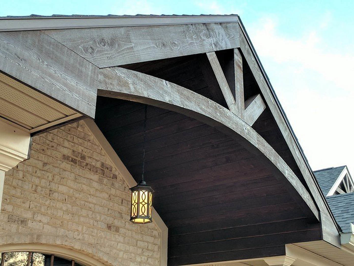 Wooden roof with exposed beams atop a brick exterior wall, outdoor light fixture mounted under the eaves, cloudy sky in background