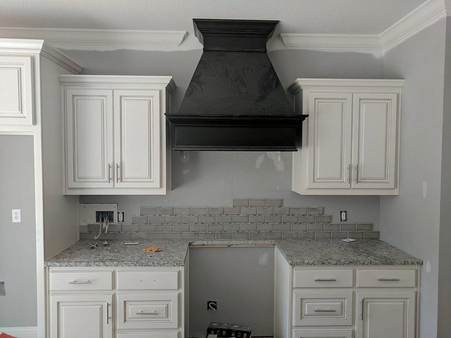 White kitchen cabinets with brushed metal handles, black vent hood mounted above a cooktop, exposed brick backsplash, white countertop, and open shelving.