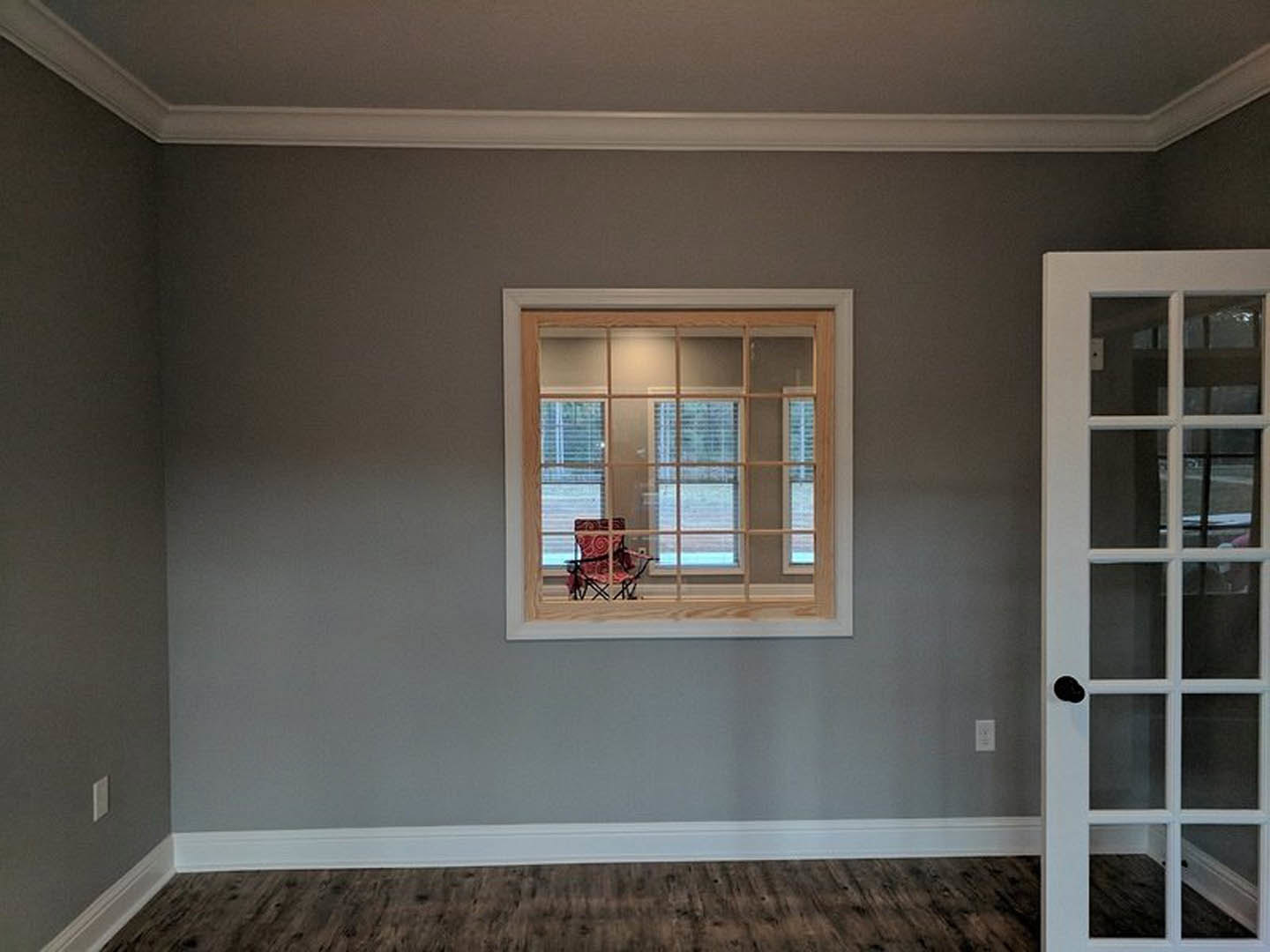 Red accent chair with white swirl pattern positioned beside a large window, white plaster walls, wood flooring, and a paneled door in a bright residential room.