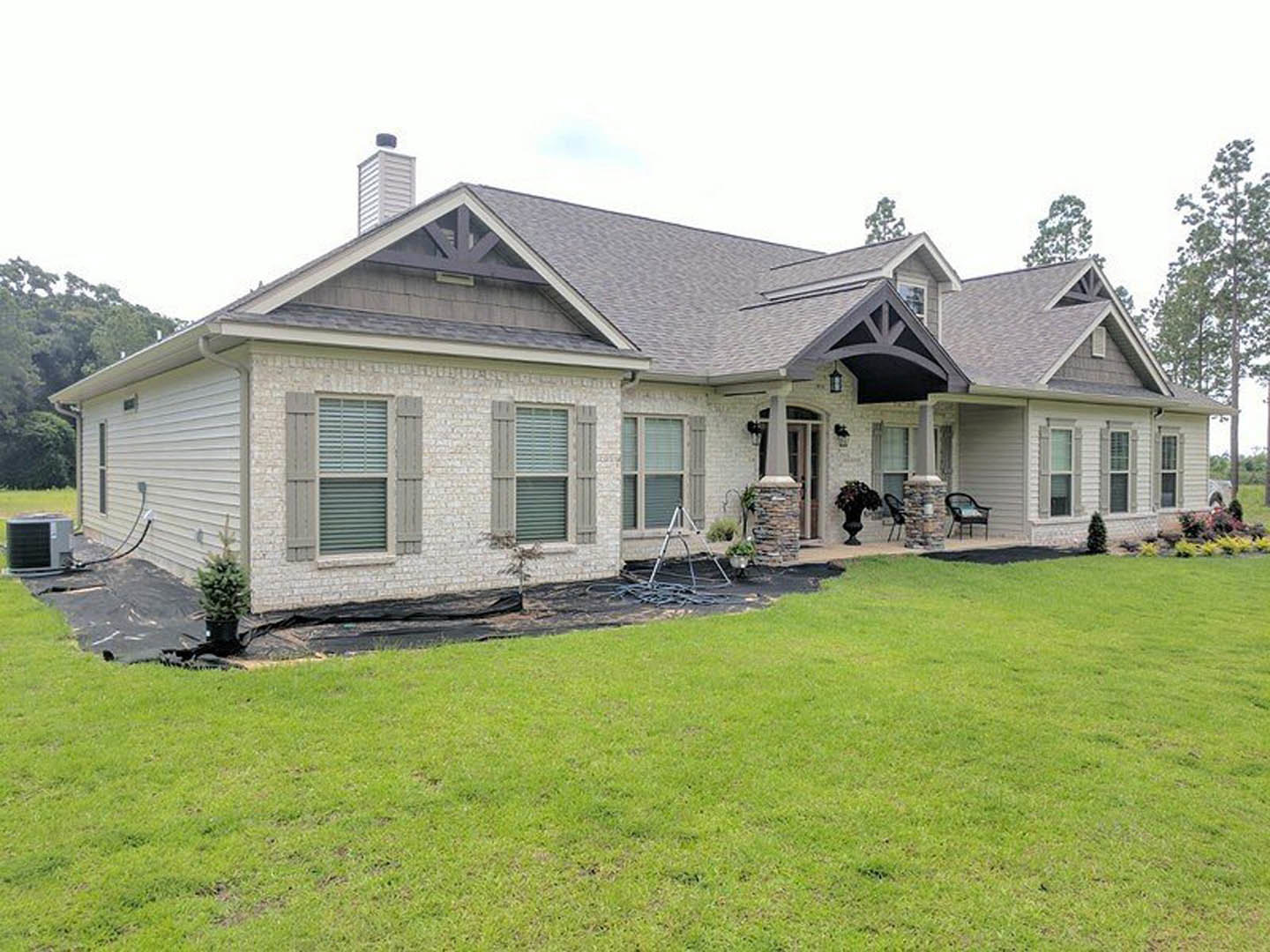 Two-story house with white siding, large windows, and a covered porch, surrounded by green lawn and mature trees; metal ladder lying on the grass near the front entrance.