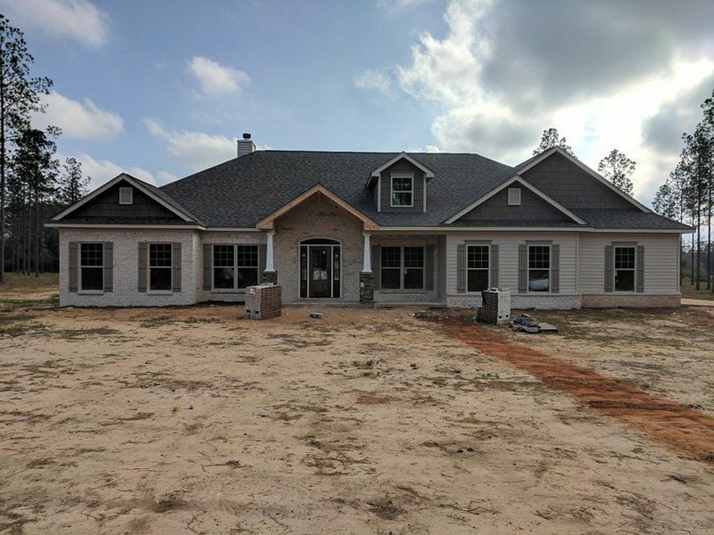 Partially built house with shingle roof, exposed framing, and dirt yard; visible windows, door opening, and stacked pallets in foreground