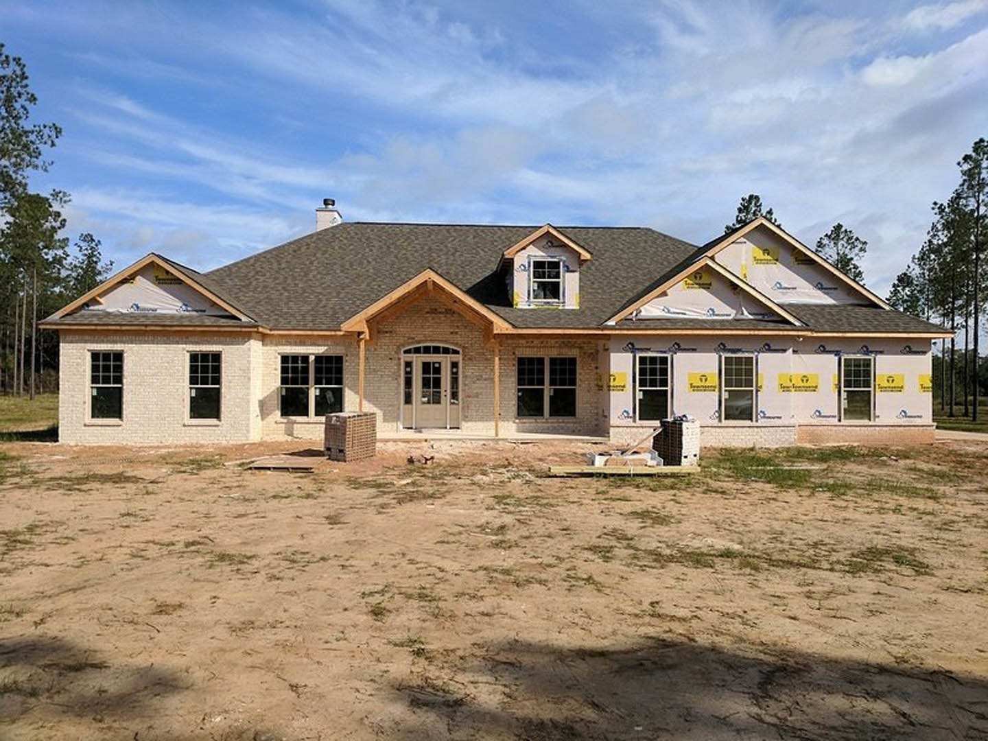 Partially built house with exposed framing and roof, surrounded by dirt field, white-framed windows visible, cloudy sky overhead