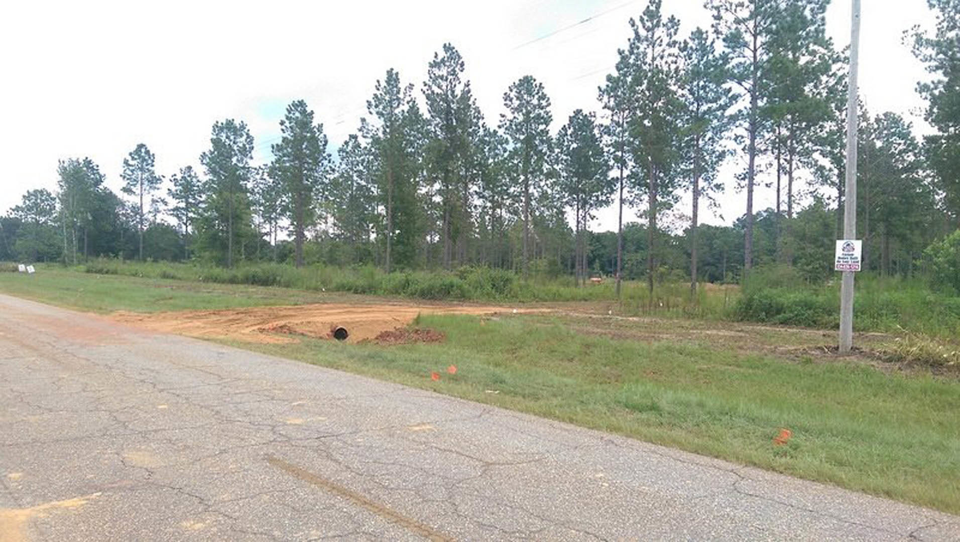 Asphalt road bordered by grass and trees, large hole in the middle of the road, sign on a pole nearby, red object partially visible in grassy area