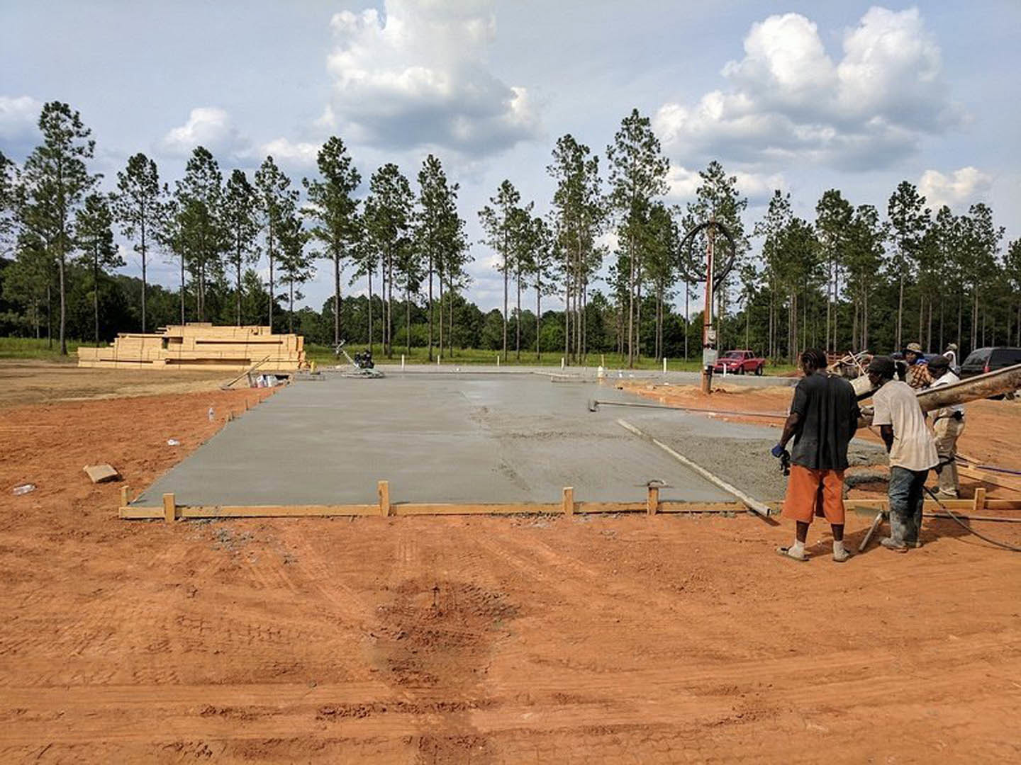 Construction site with several men standing on a dirt surface near a cement slab, surrounded by trees and open sky